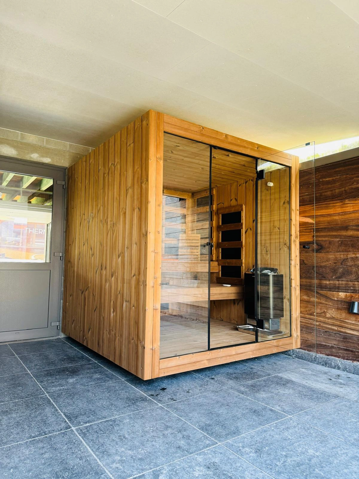 Luxe kubusvormige design buitensauna met glazen voorwand en moderne thermowood afwerking, in de tuin voor een ultieme outdoor wellness ervaring.