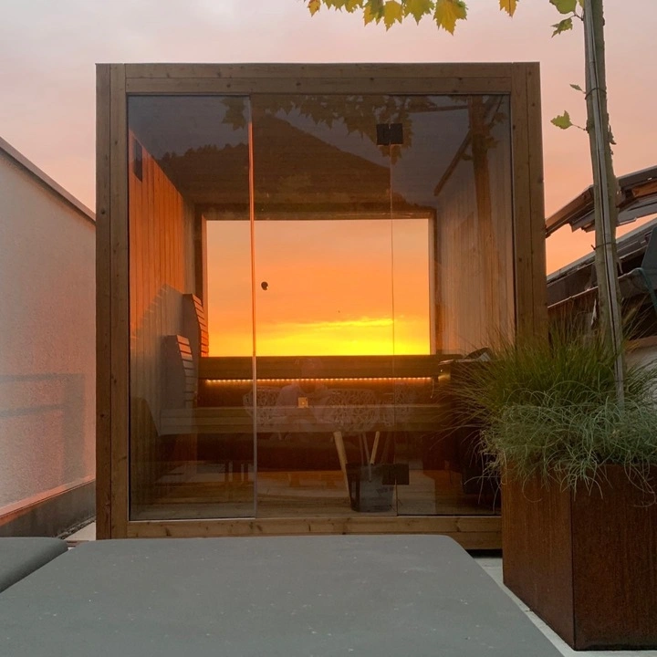 Luxe kubusvormige design buitensauna met glazen panorama achterwand, glazen voorwand en moderne thermowood afwerking, in de tuin voor een ultieme outdoor wellness ervaring.