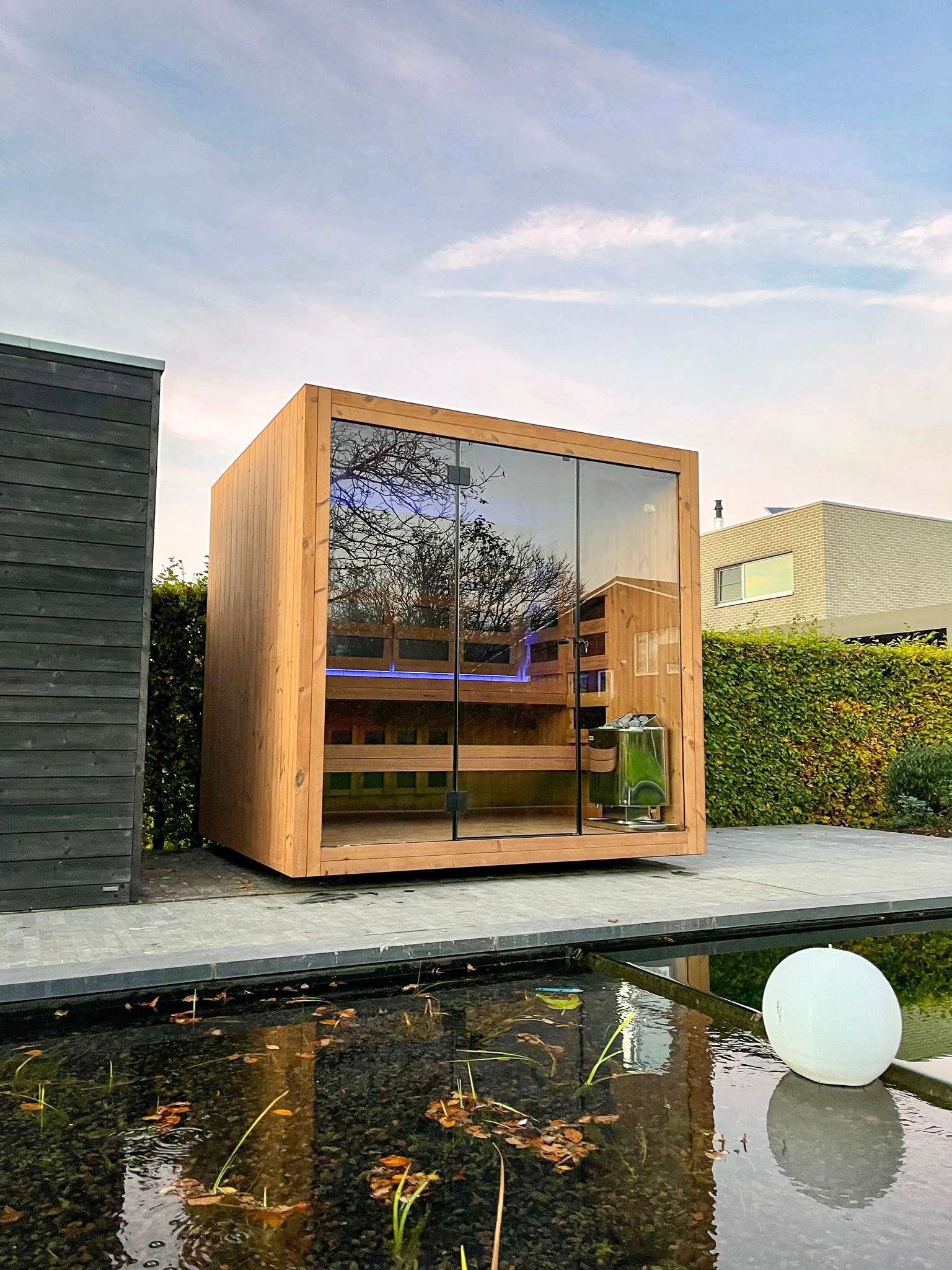 Luxe kubusvormige design buitensauna met infrarood glazen voorwand en moderne thermowood afwerking, in de tuin voor een ultieme outdoor wellness ervaring.