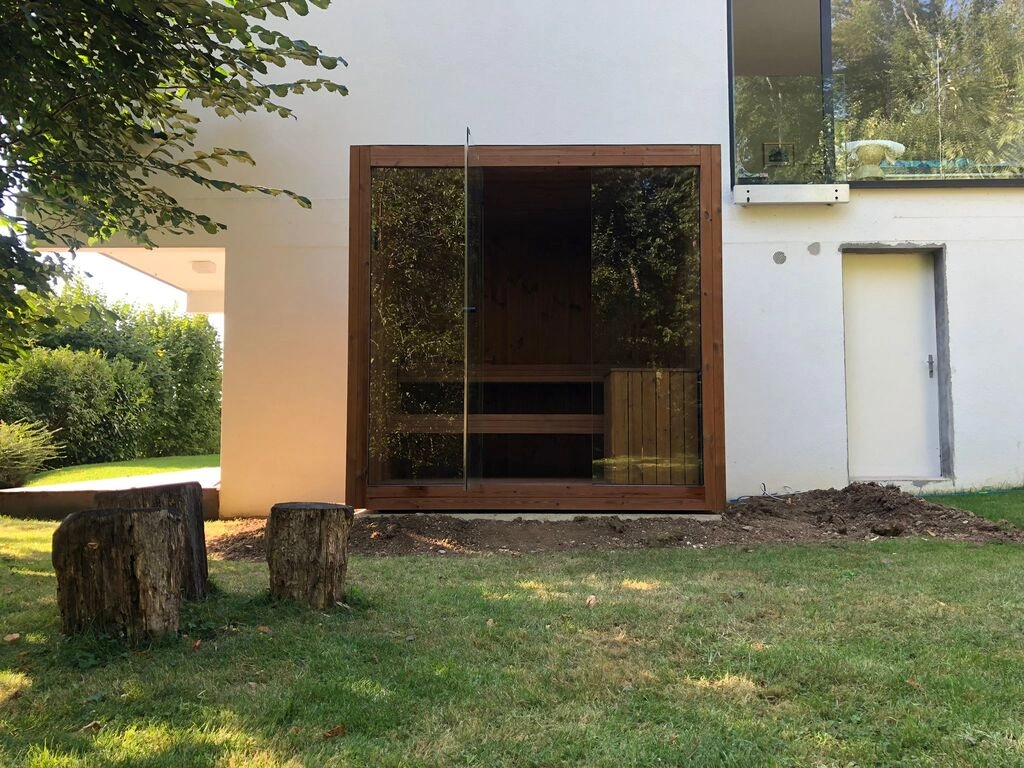 Luxe kubusvormige design buitensauna met glazen voorwand en moderne thermowood afwerking, in de tuin voor een ultieme outdoor wellness ervaring.