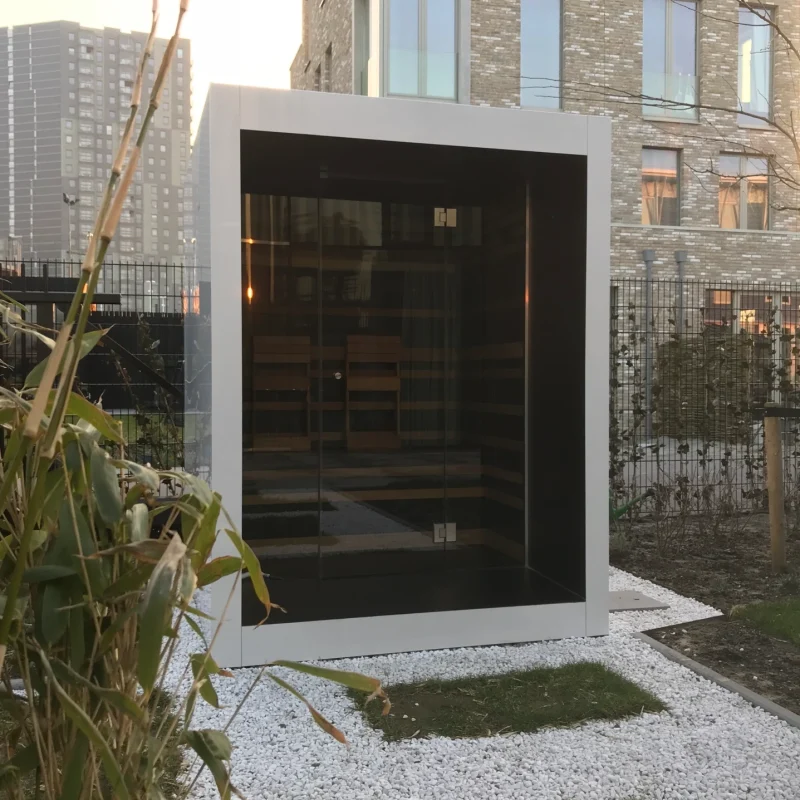 Compact luxury cube-shaped design outdoor sauna with glass front and modern inox finish, in the garden for the ultimate outdoor wellness experience.