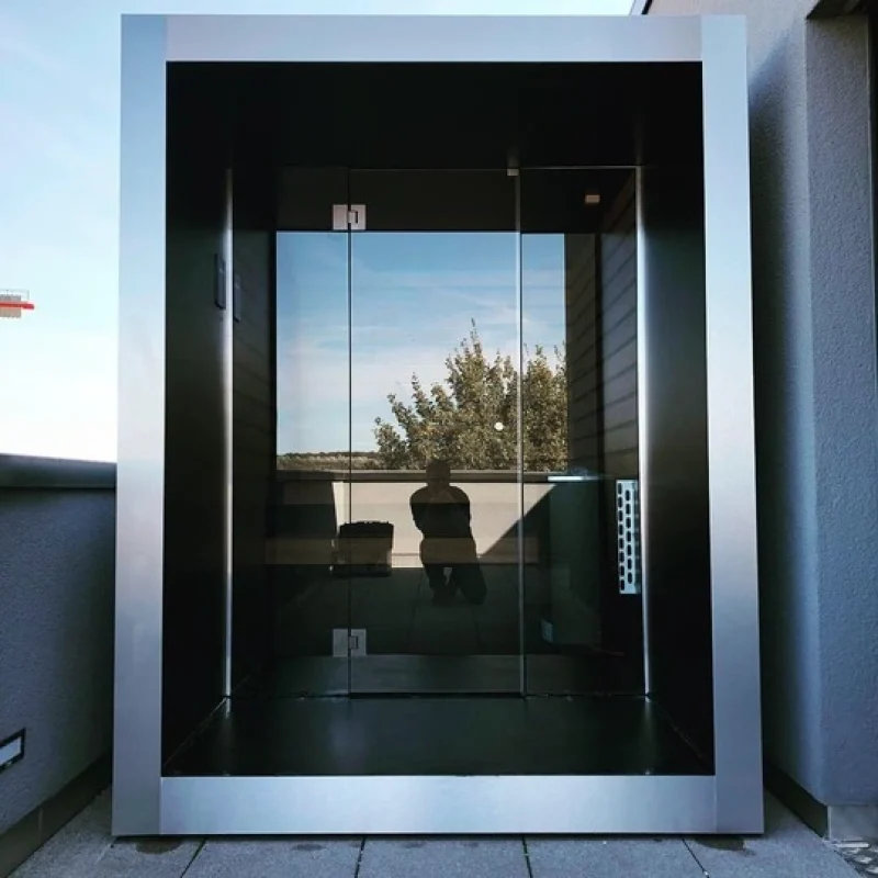 Compact luxury cube-shaped design outdoor sauna with glass front and modern inox finish, in the garden for the ultimate outdoor wellness experience.